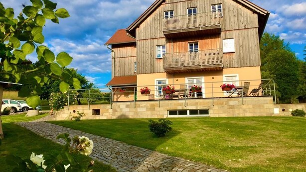 A multi-story wooden building with balconies, a paved driveway, and a well-maintained lawn. - Ferienwohnung Winterstein (Sebnitz)
