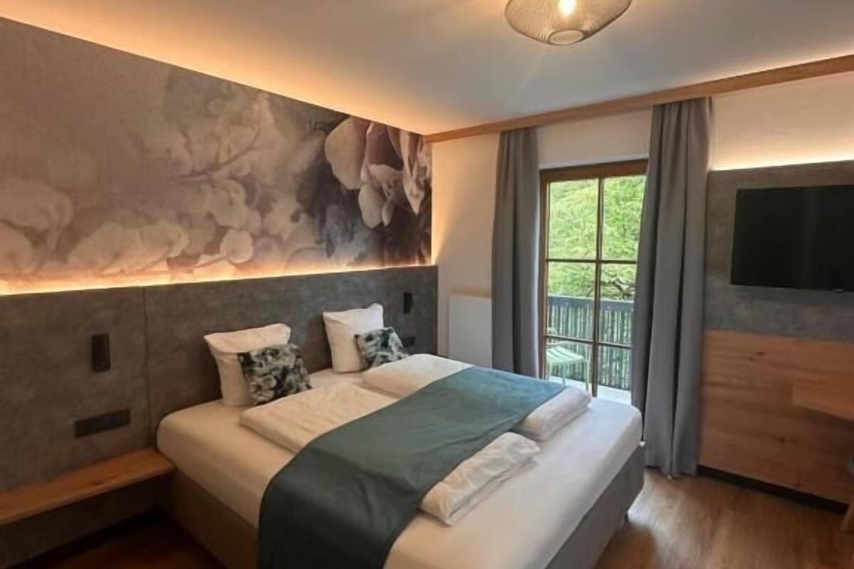 Une chambre moderne avec un grand lit, un téléviseur fixé au mur et une vue sur la verdure par la fenêtre.