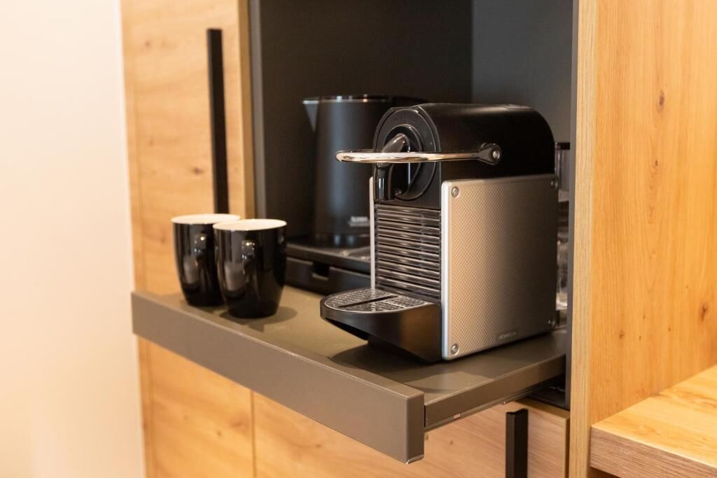 A compact kitchenette with a coffee machine, two black mugs, and a wooden cabinet.