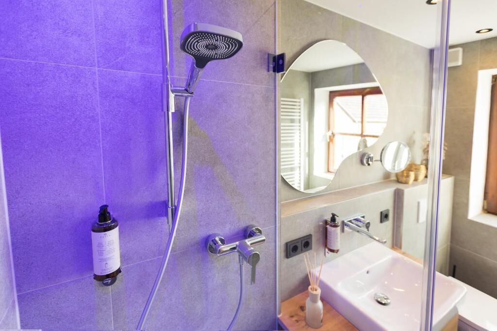 A modern bathroom with a purple-tiled wall, a handheld showerhead, a sink with a mirror, and a bottle of shampoo.