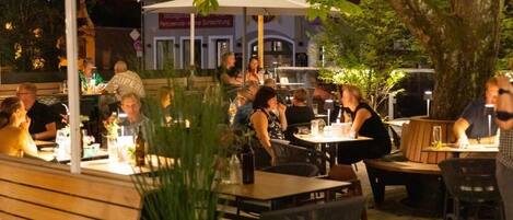 An outdoor dining area with tables and chairs, surrounded by greenery and umbrellas.