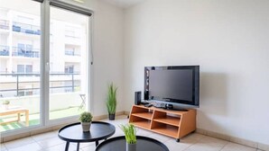 Una sala de estar moderna con un televisor grande sobre un mueble de madera, una pequeña mesa redonda con una planta y un ventanal corredero que da a un balcón.