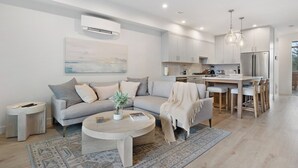A modern living room with a grey sofa, a coffee table, and a kitchen area with white cabinets and a stainless steel refrigerator.