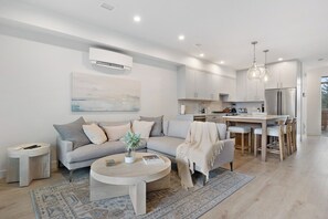A modern living room with a grey sofa, a coffee table, and a kitchen area with white cabinets and a stainless steel refrigerator.