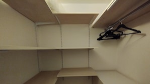Et hjørne af et rum med beige vægge og et hvidt loft. Tre hvide hylder sidder fastgjort til væggen, og en sort bøjle hænger på højre side.