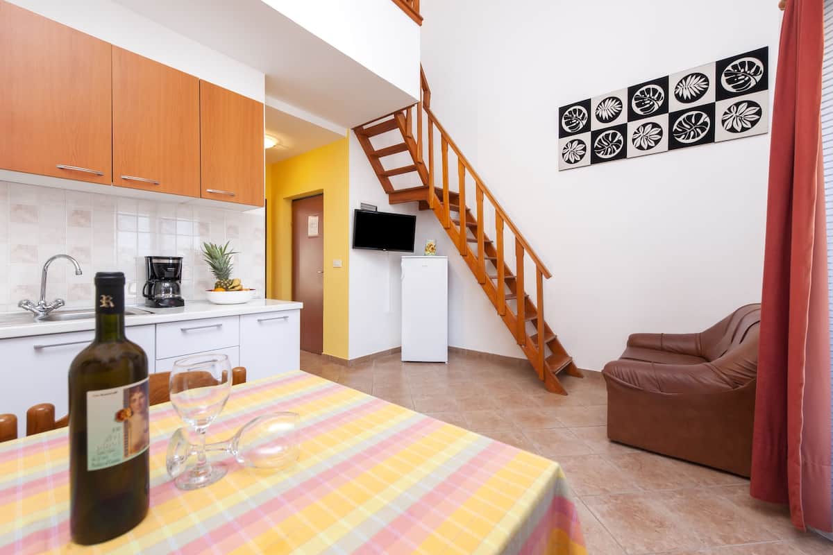 A modern kitchen with wooden cabinets, a dining table with a checkered tablecloth, and a staircase leading to a living area with a sofa and a TV.