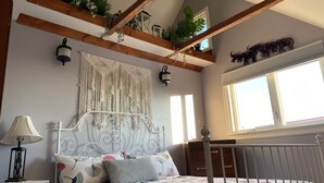A bedroom with a bed, bedside table, lamp, macrame wall hanging, wooden beams, and a window. - Relax on the South Shore, steps from 2 beaches, ocean views & hot tub. (East River Point)