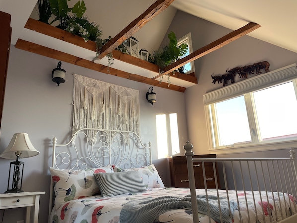 A bedroom with a bed, bedside table, lamp, macrame wall hanging, wooden beams, and a window.