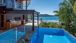 Un espace piscine avec vue sur l’océan, une terrasse en bois et une cuisine extérieure.