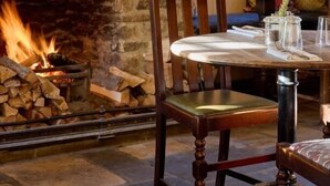 A dining area with a stone fireplace, a wooden chair, and a table set with a glass pitcher and a plate. - The Plough Inn (Lechlade)