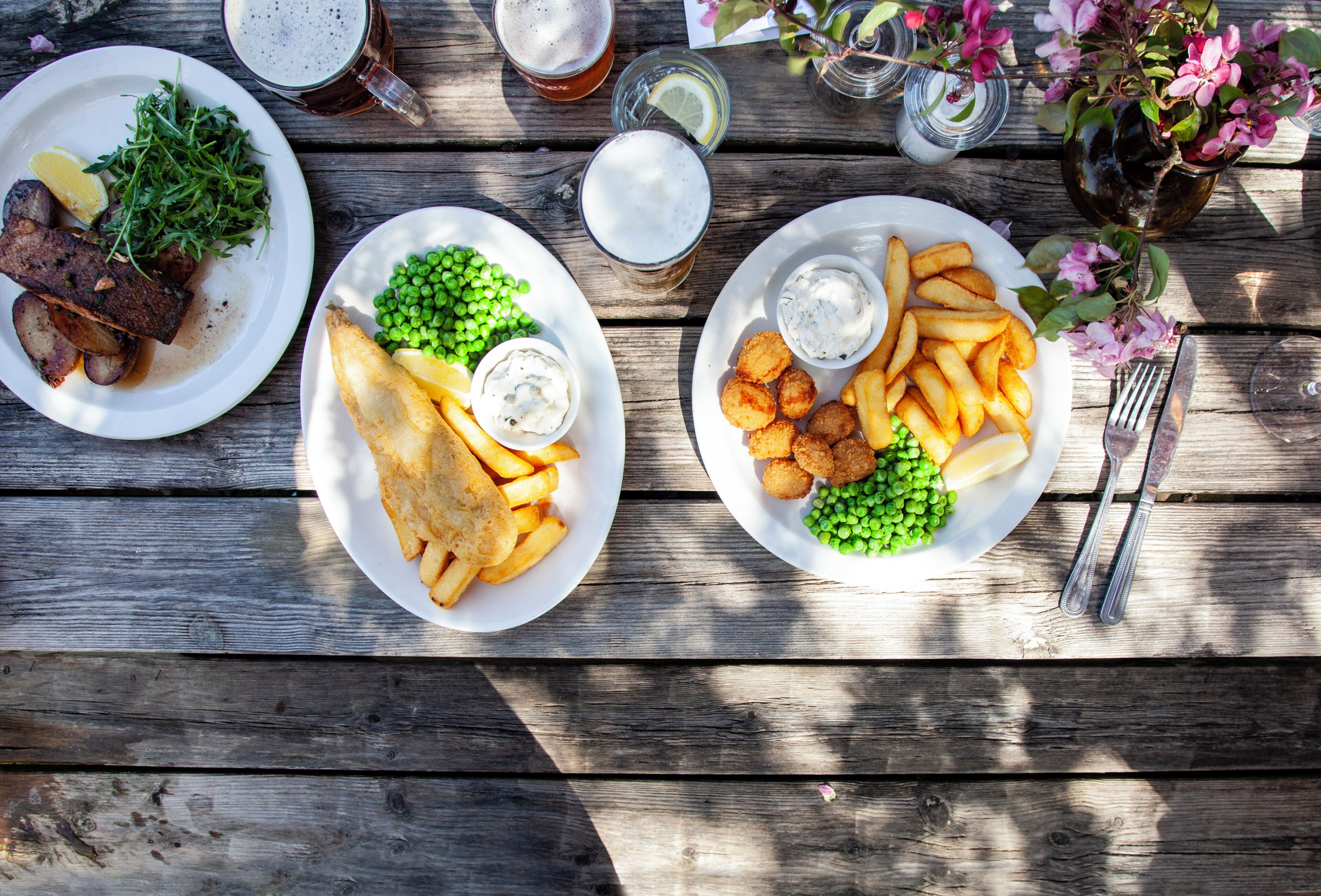 A wooden table with two plates of food, a glass of beer, and a vase with flowers.
