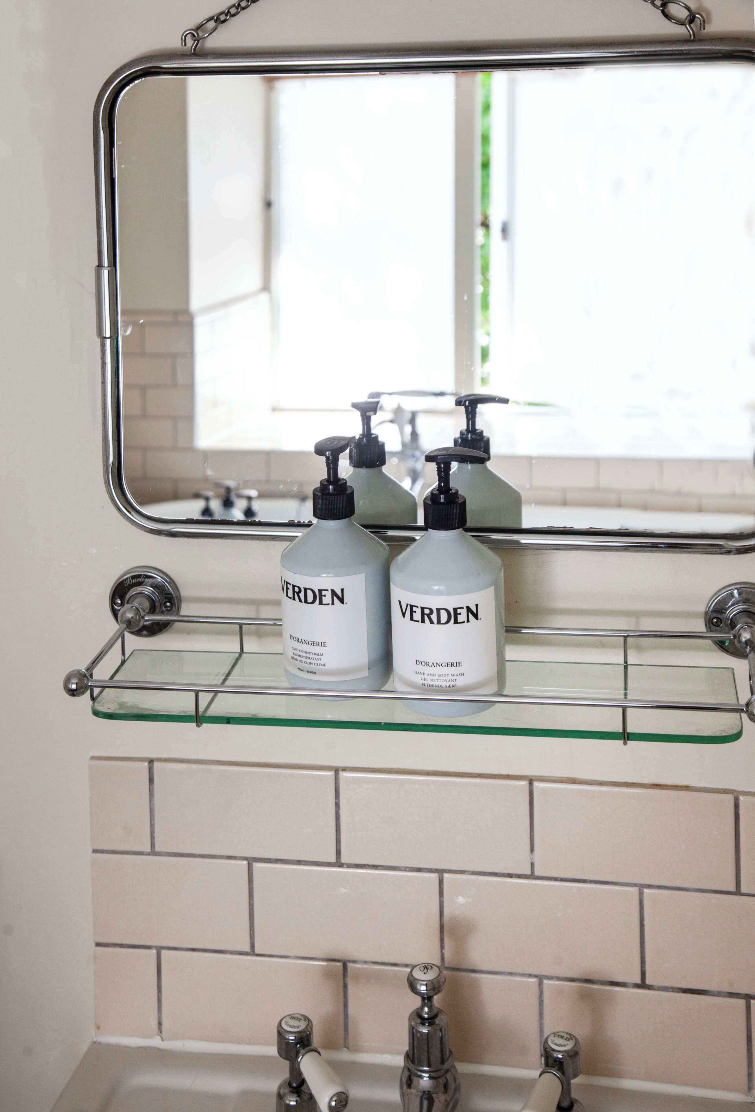 Un bagno con ripiano in vetro che contiene due flaconi di bagnoschiuma e crema corpo Verden, un rubinetto cromato e uno specchio sopra.