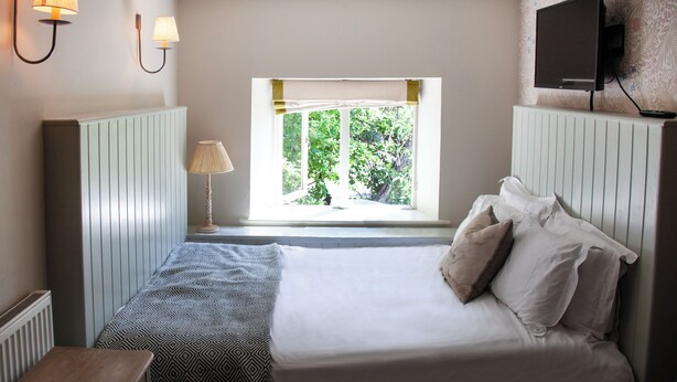 A bedroom with a bed, a window with a view of greenery, a bedside table, and a lamp. - The Plough Inn (Lechlade)