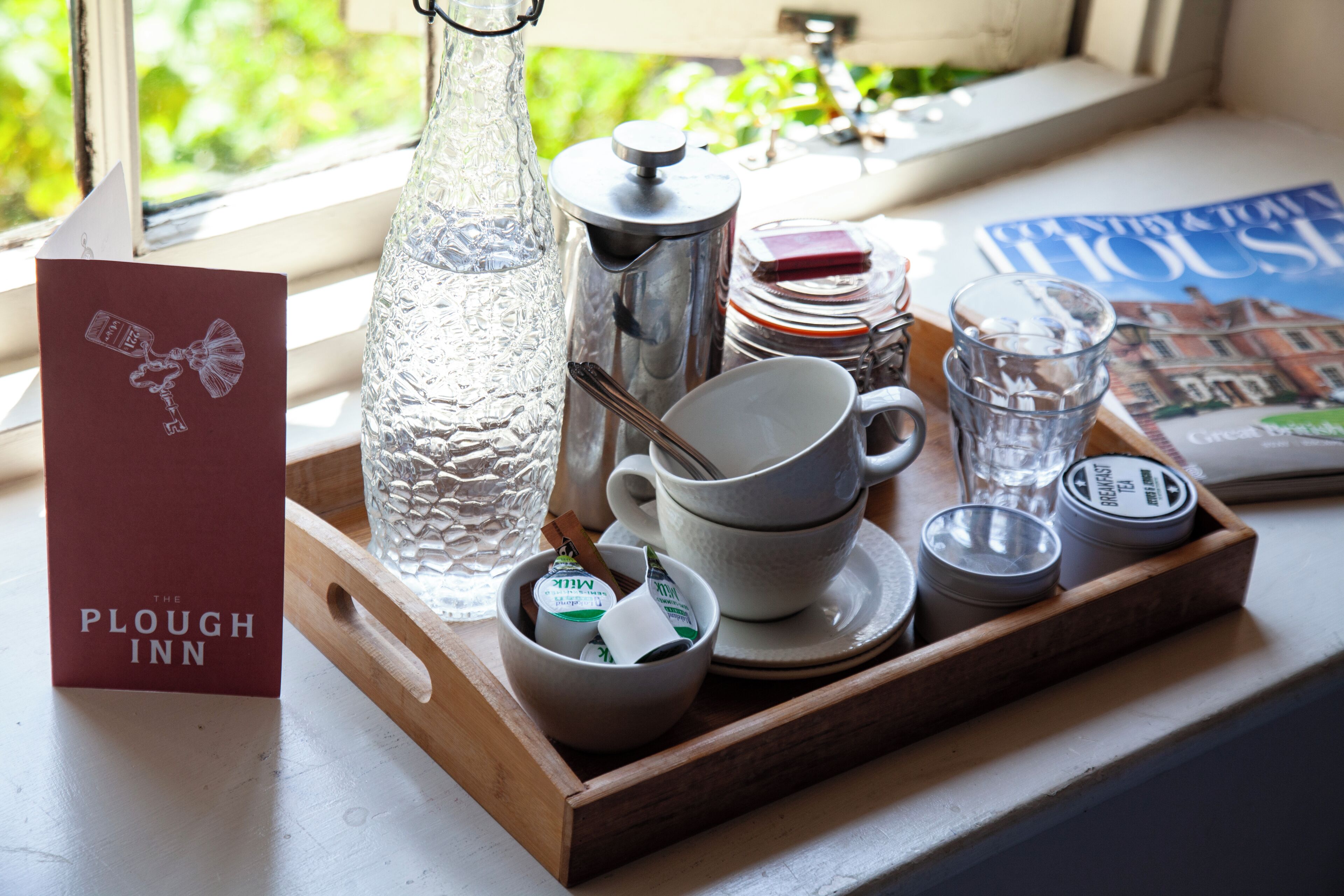 Bandeja de desayuno con cafetera, leche, té y una tarjeta sobre una mesa de madera junto a una ventana.