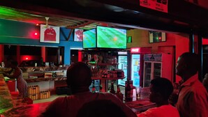 Un bar con múltiples pantallas mostrando partidos deportivos, un refrigerador de Coca-Cola y una variedad de botellas y vasos sobre el mostrador.