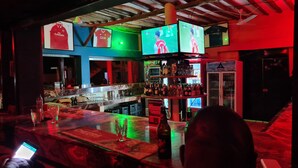 Zona de bar con varias pantallas mostrando contenido deportivo, barra con botellas y vasos, y techo con vigas y luces a la vista.