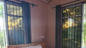 Una cama individual con estructura de madera y colchón blanco, una mesita pequeña y una ventana con barrotes y cortinas.