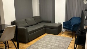 Un salon moderne avec un canapé d’angle, une petite table, un fauteuil, un téléviseur et une horloge au mur.