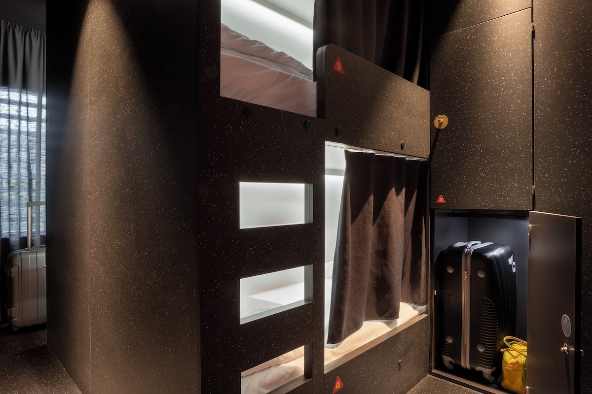 Habitación compacta con cama, un ventanal con cortinas, espacio para guardar equipaje y un calentador fijado a la pared.