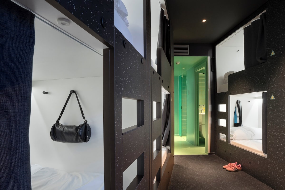 Un pasillo estrecho con literas, un bolso colgado y un baño iluminado con luz verde.