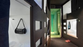 A narrow hallway with bunk beds, a hanging bag, and a green-lit bathroom.