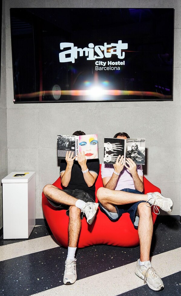 Deux personnes sont assises sur un fauteuil en forme de sac de haricots rouge, devant un mur sur lequel est affiché un panneau indiquant « Amistat City Hostel Barcelona ».