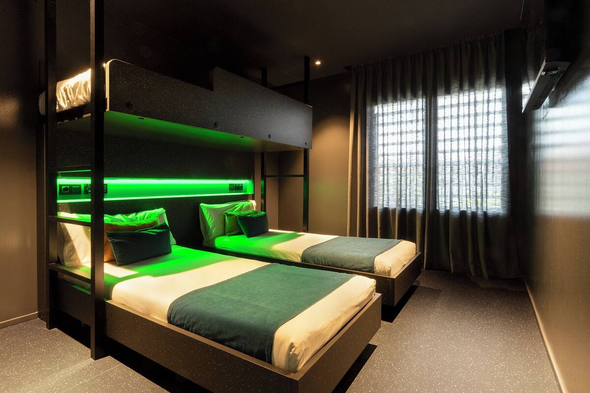 Una habitación de hotel moderna con literas, un cabecero iluminado en verde y un amplio ventanal con cortinas.