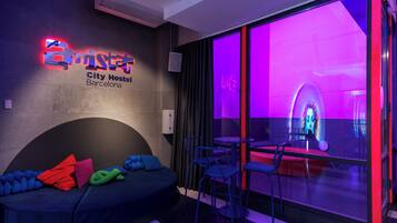 Un dormitorio moderno de hostal con un letrero de neón, un sofá con cojines de colores y un amplio ventanal con vistas a la ciudad.