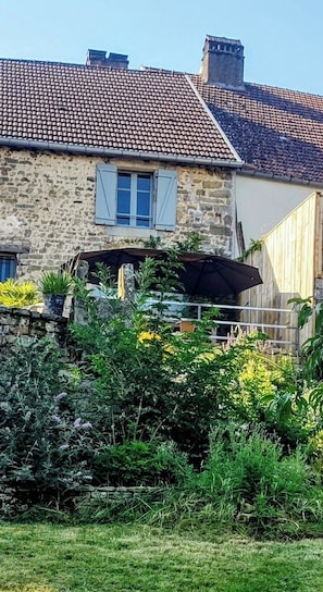 Une maison en pierre avec un toit en tuiles, une fenĂȘtre avec des volets et un jardin oĂč poussent diffĂ©rentes plantes.