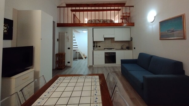 Sala de estar com televisão, sofá azul, mesa de centro com azulejos e área de cozinha com armários e fogão.