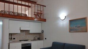 Cozinha compacta com armários brancos, fogão e pia. Há um sofá azul e um quadro na parede.