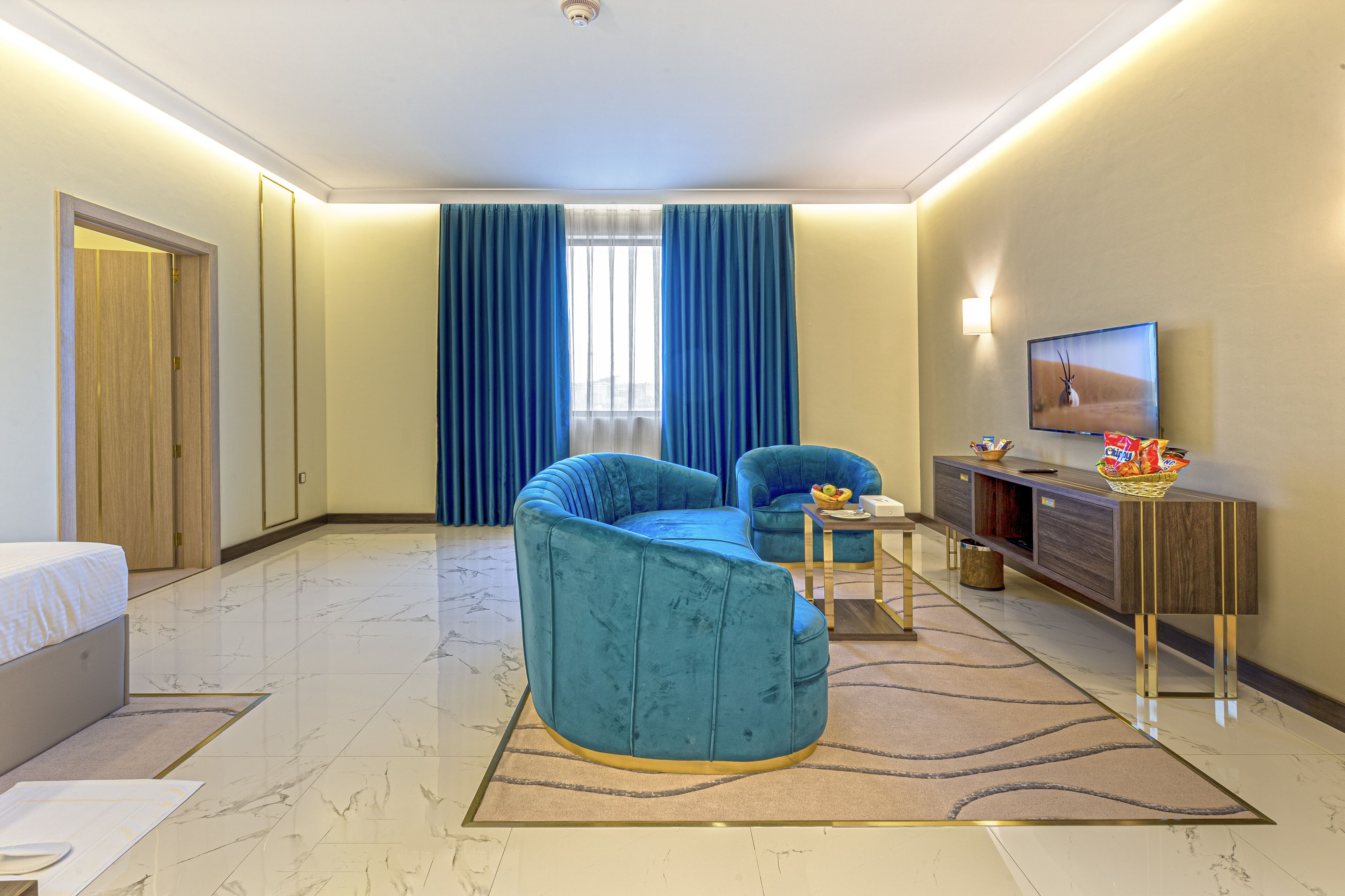 A modern hotel room with a curved blue sofa, a flat-screen TV, a wooden cabinet, and a large window with blue curtains.