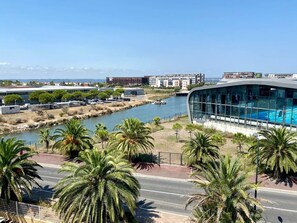 A modern building with a glass facade overlooking a body of water, palm trees, and a road. - Appartement 4e Étage 300 m Plage (Sète)