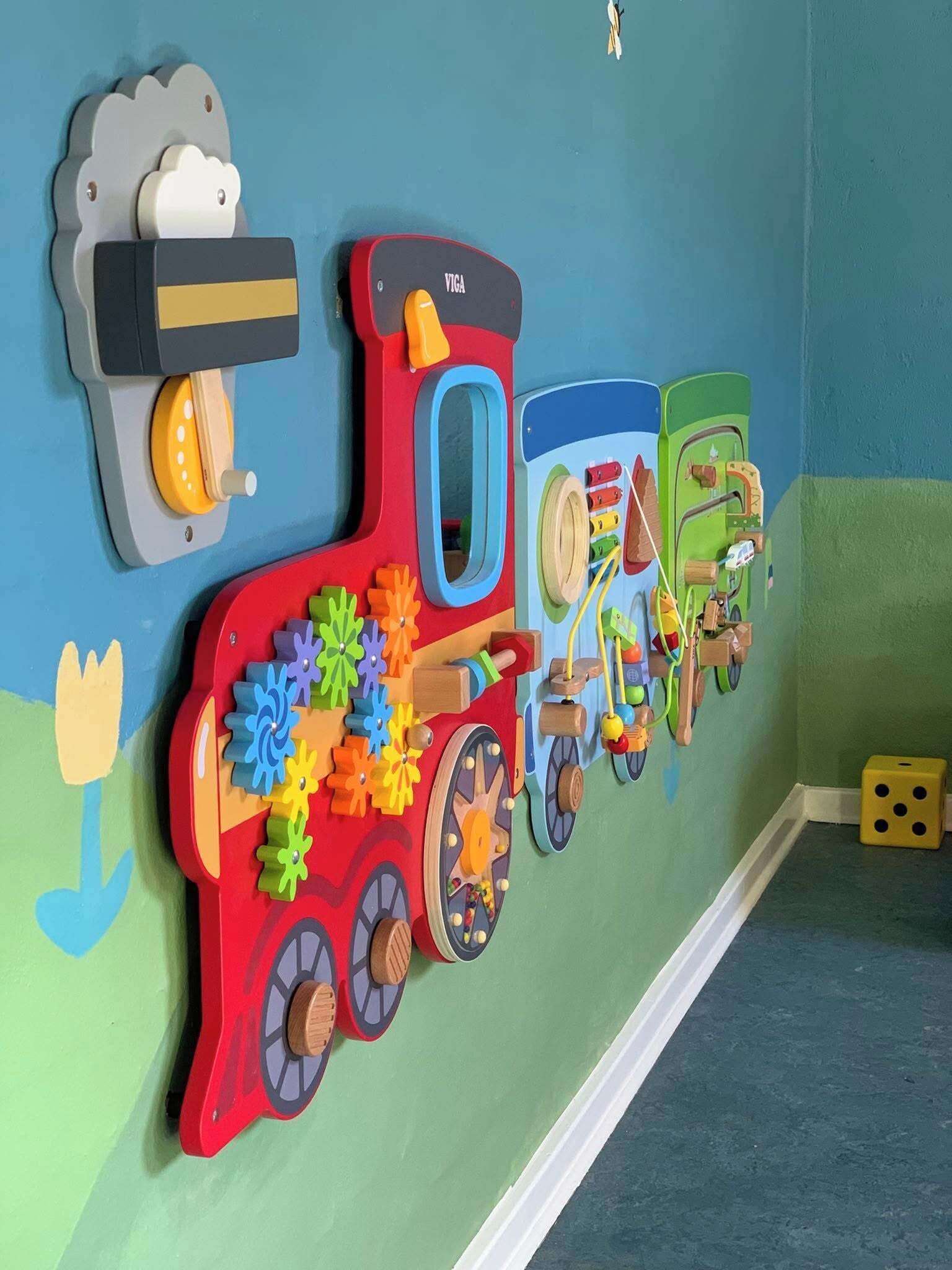 Un tren de juguete infantil colorido con ruedas y engranajes, montado en una pared con fondo azul y verde.