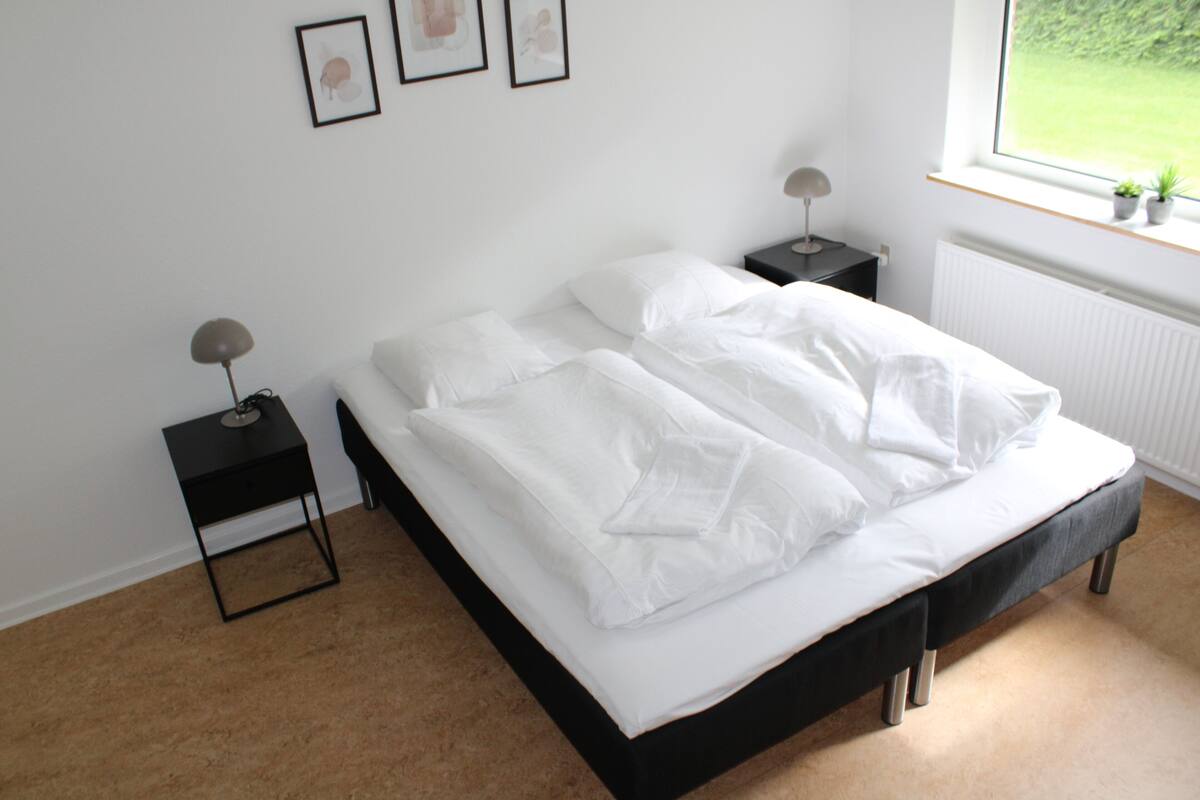 Ein Schlafzimmer mit einem Bett, zwei Nachttischen, einem Fenster mit Blick ins Grüne und gerahmten Kunstwerken an der Wand.