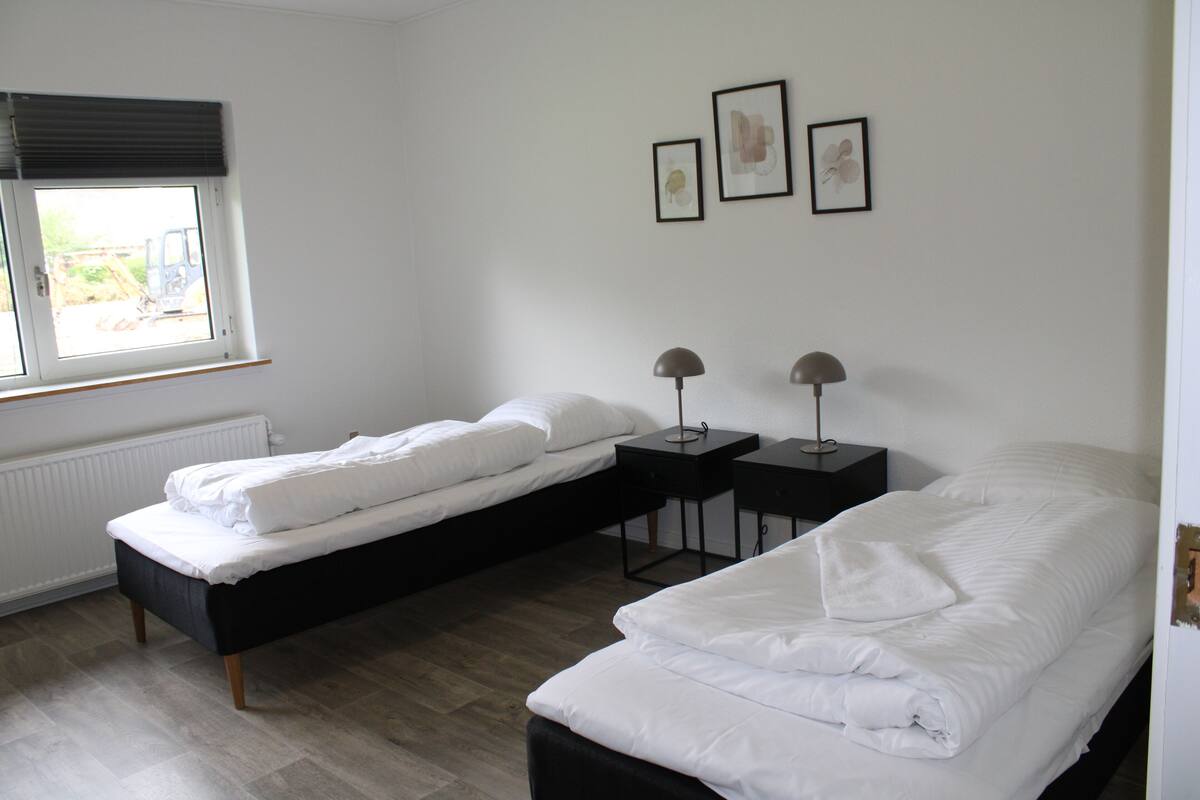 Ein Schlafzimmer mit zwei Einzelbetten, einem Fenster mit Jalousien, einem Heizkörper und drei gerahmten Bildern an der Wand.