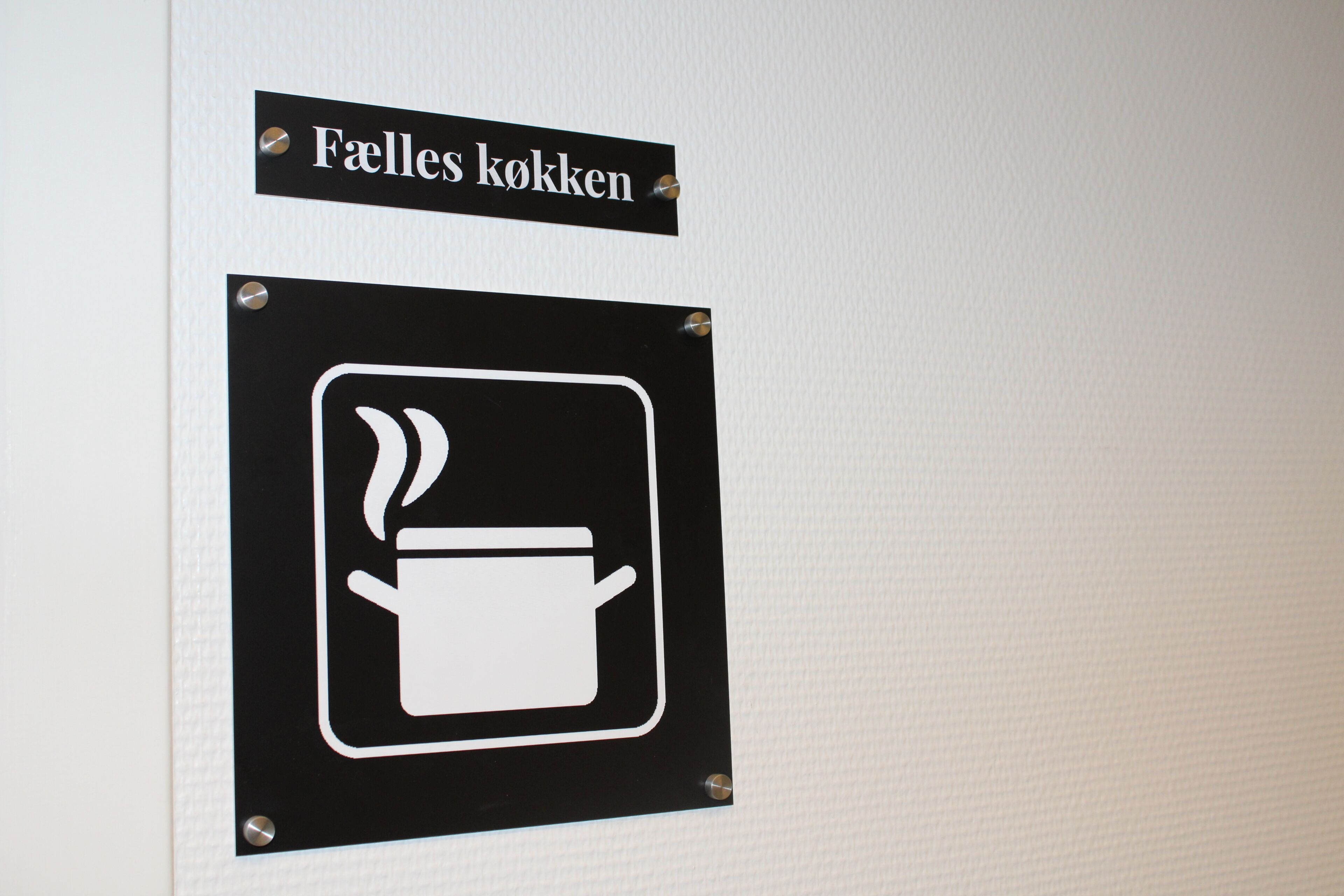 A sign with a coffee pot symbol and the text 'Faelles kökken' indicating a coffee area.