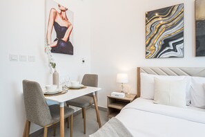Habitación de hotel con cama, una mesa de comedor con sillas, una mesita de noche con lámpara y dos obras de arte en las paredes.