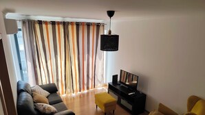 Un salon comprenant un canapé, un pouf et un fauteuil, une télévision et un luminaire suspendu noir.