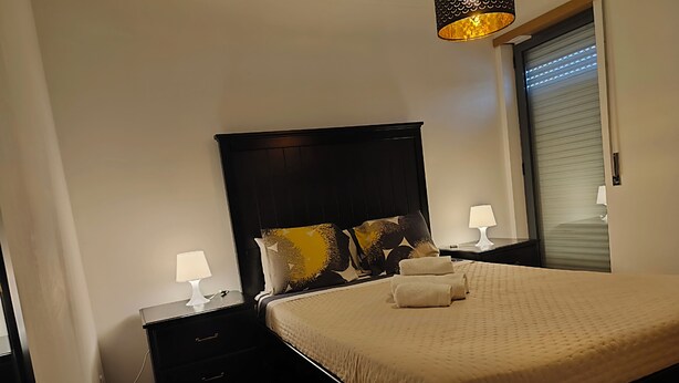 Un lit bien fait, avec une couette beige, deux tables de chevet avec des lampes, une tête de lit noire et une fenêtre avec des stores.