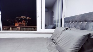 Een slaapkamer met een groot raam, een bed met grijze beddengoed en uitzicht op een gebouw 's nachts.