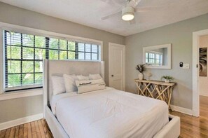 A bedroom with a large bed, a ceiling fan, a window with blinds, a bedside table with a mirror, and a door.