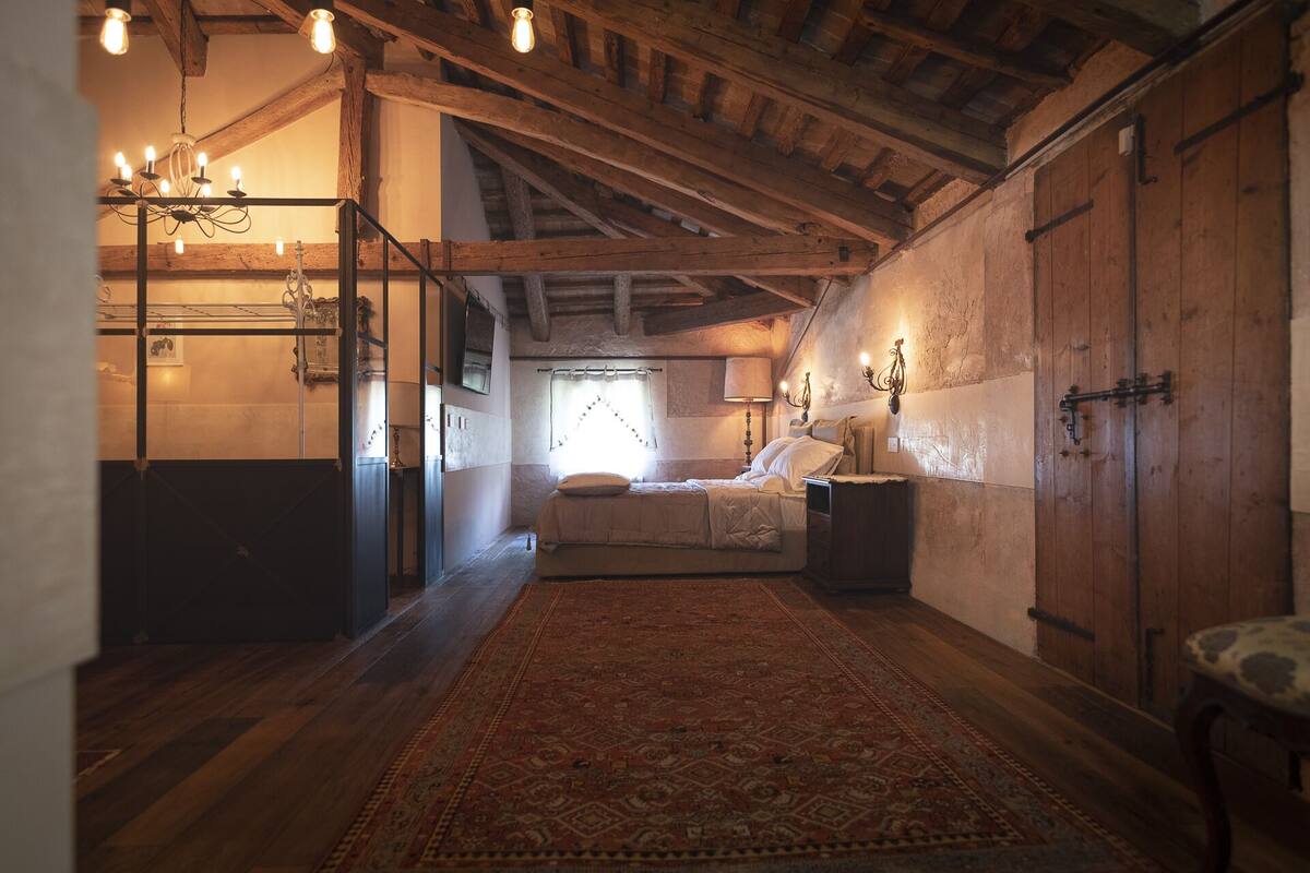 A rustic bedroom with exposed wooden beams, a bed with a red patterned rug, a chandelier, and a wooden wardrobe.