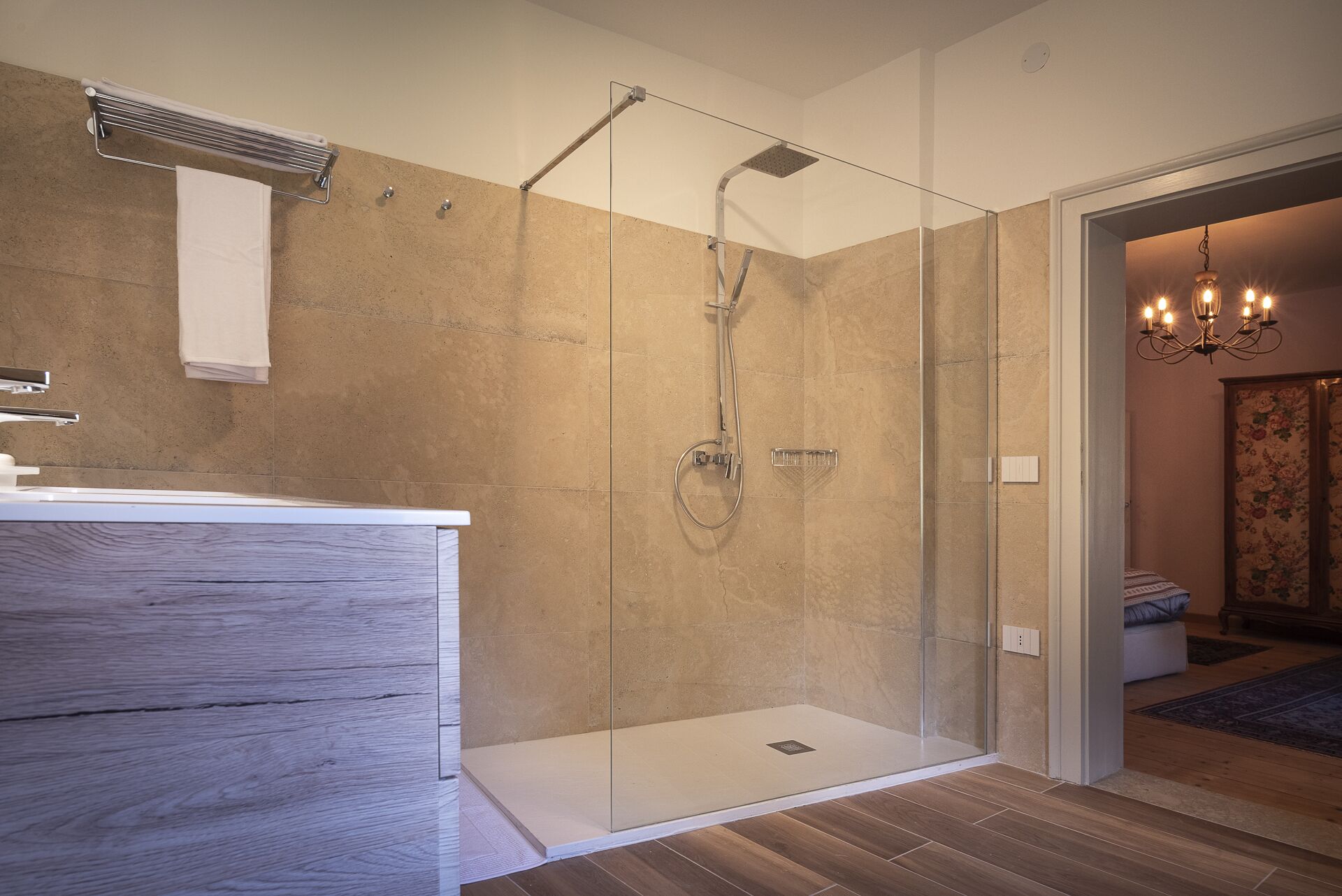 A modern bathroom with a glass-enclosed shower, a wooden vanity, and a chandelier in the adjacent room.
