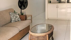 Una sala de estar con un sofá, una mesa de centro de vidrio, un ventilador negro y una zona de cocina con armarios blancos y un ventilador de techo.