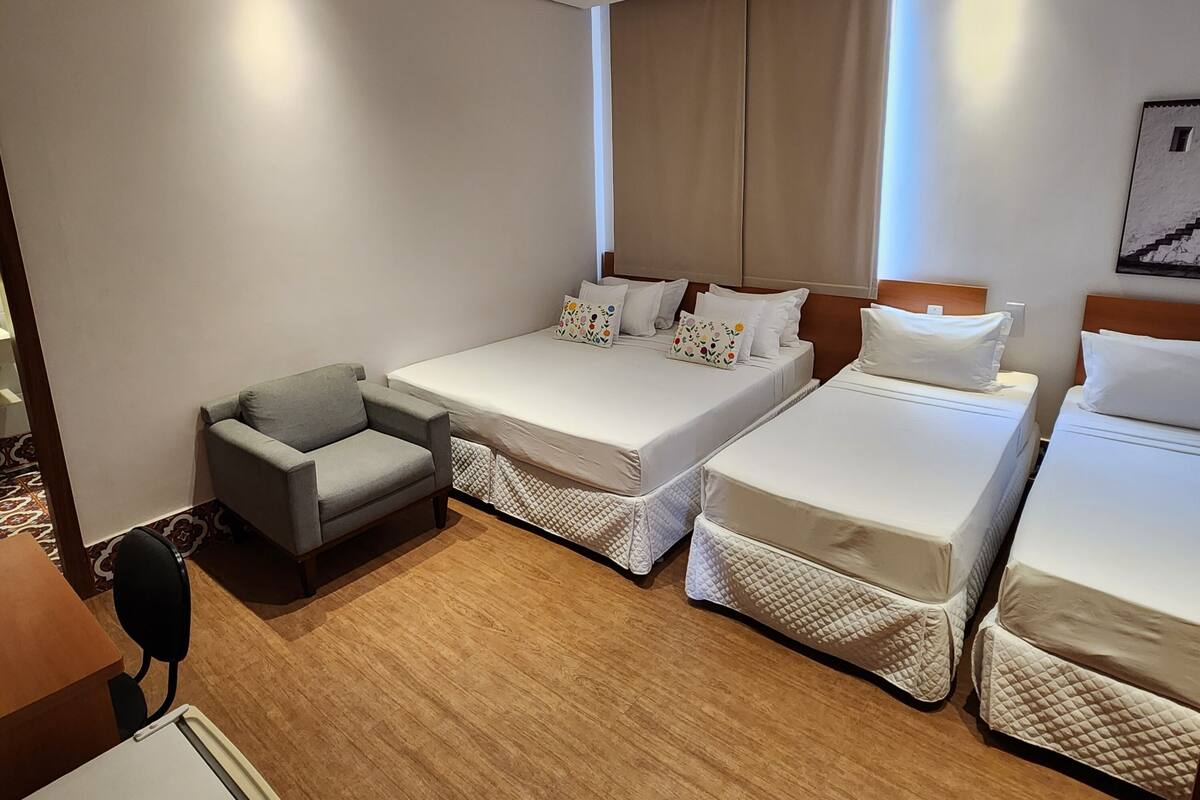 Une chambre d’hôtel avec deux lits simples, un fauteuil gris, un plancher en bois, un ventilateur de plafond et une fenêtre avec des rideaux.