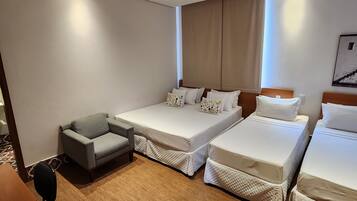 A hotel room with two single beds, a gray armchair, a wooden floor, a ceiling fan, and a window with curtains.