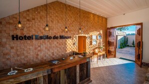 Een receptiehal met een bakstenen muur, een houten receptiebalie, hangende pendantlampen en een bordje met de tekst 'Hotel Estilo de Minas'.