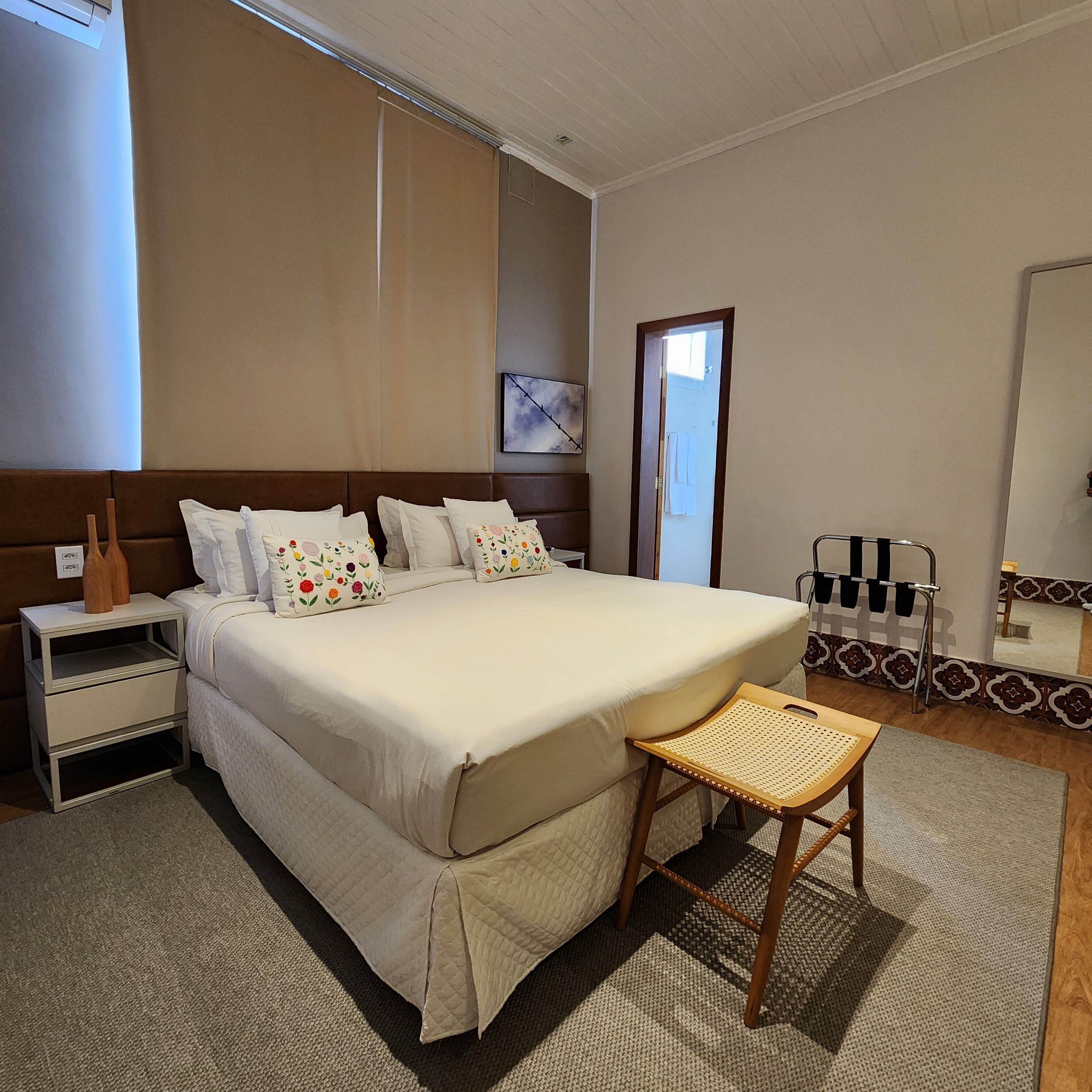 Un dormitorio ordenado con una cama grande, una mesita de noche, una silla y un espejo.
