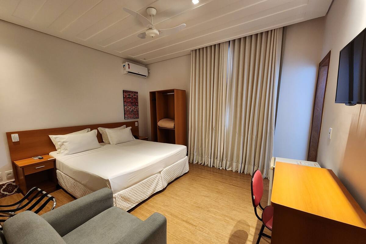 Une chambre d’hôtel comprenant un lit, un bureau, une chaise, une armoire, un ventilateur de plafond et une fenêtre avec des rideaux.